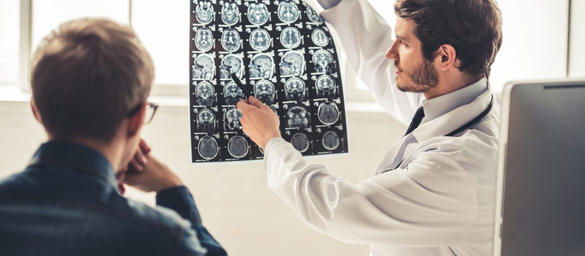 Handsome young doctor in white coat is showing x-ray image to his patient while working in office